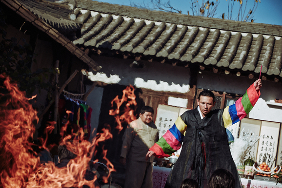 Un hombre lleva puesto un hanbok colorido y hay llamas frente a una casa tradicional coreana (hanok).
