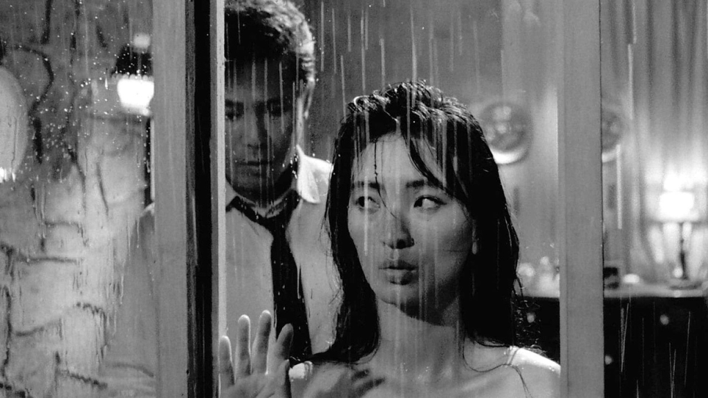 Foto en blanco y negro: llueve fuera de la ventana, y detrás de la mujer se ve a un hombre.
